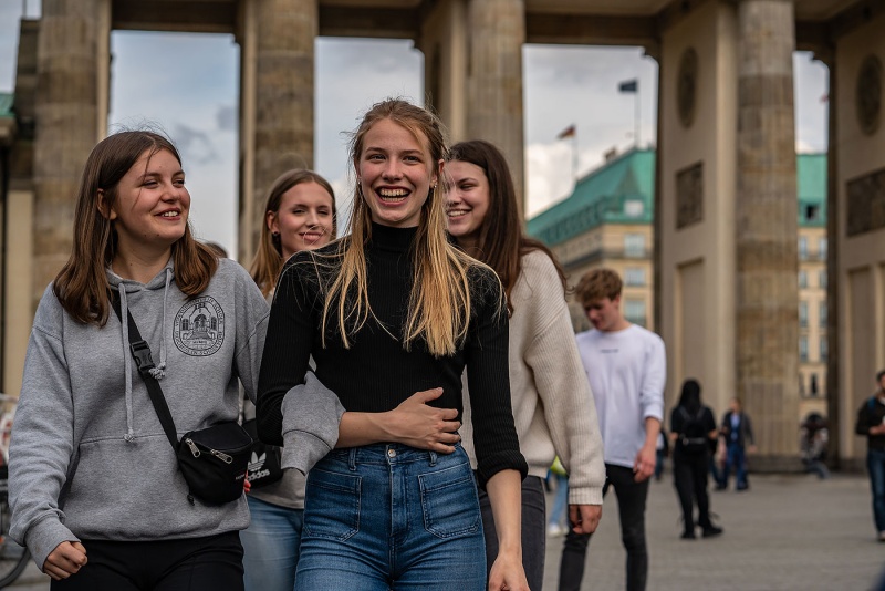 Vier Schülerinnen laufen lachend durch Berlin. Im Hintergrund das Brandenburger Tor und das Hotel Adlon.