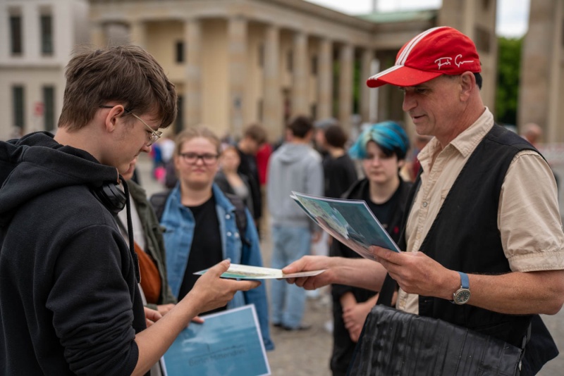 Ein Stadtführer gibt einem Schüler vor dem Brandenburger Tor eine Karte mit Aufgaben für Rallye durch Berlin.